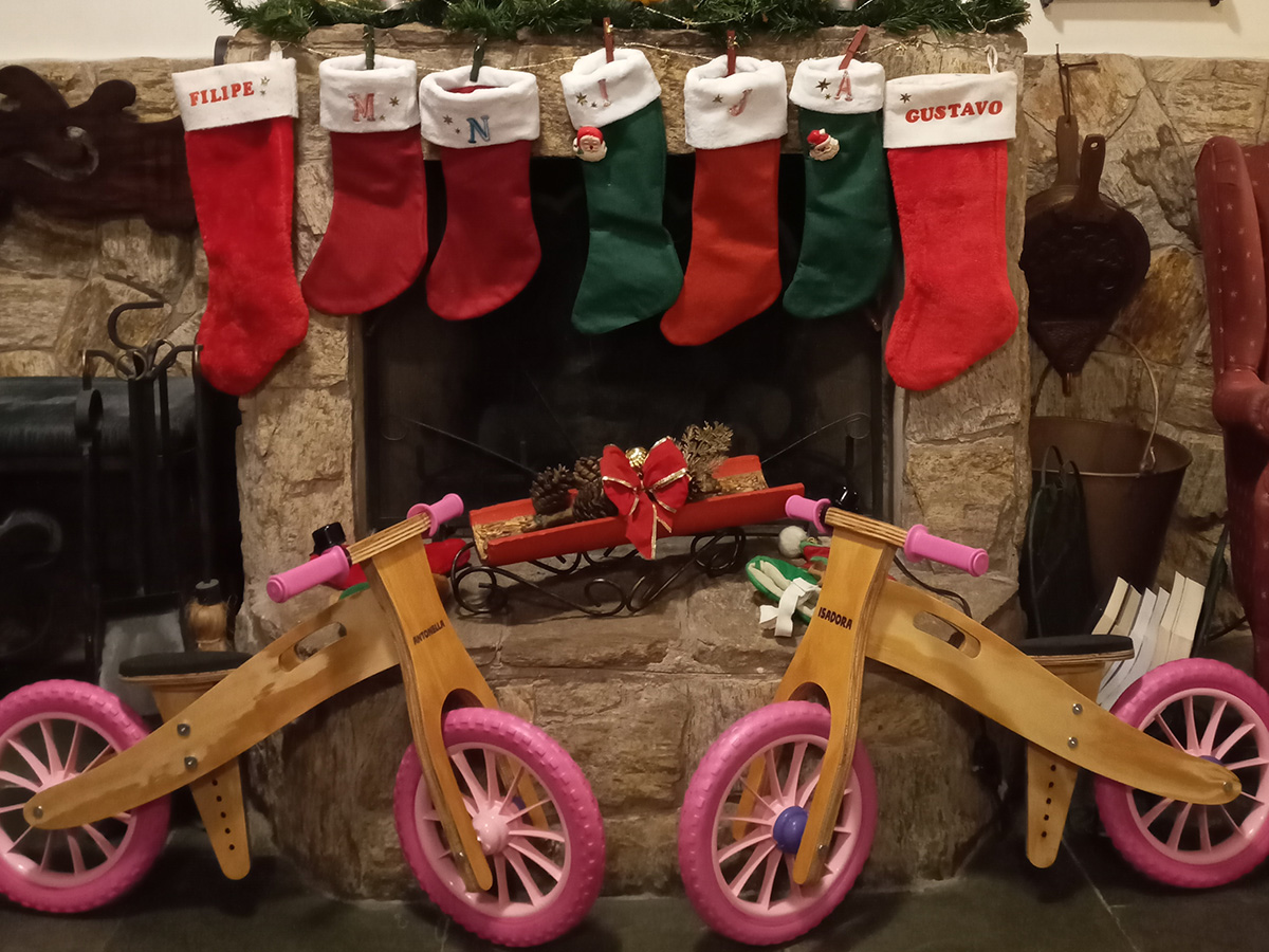 Duas Biciquetinhas de madeira com rodas rosa e os nomes Antonella e Isadora gravados no quadro, em frente à lareira decorada com botas de Natal